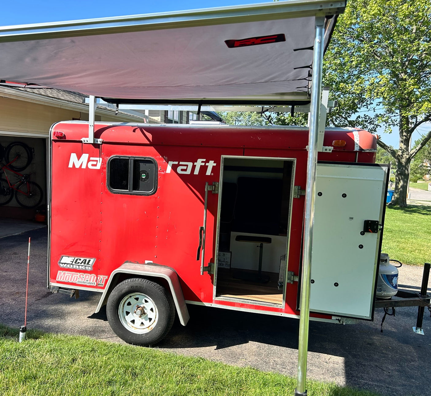 Red enclosed trailer with an awning and open side door, appearing as a compact RV rental. Features a propane tank.