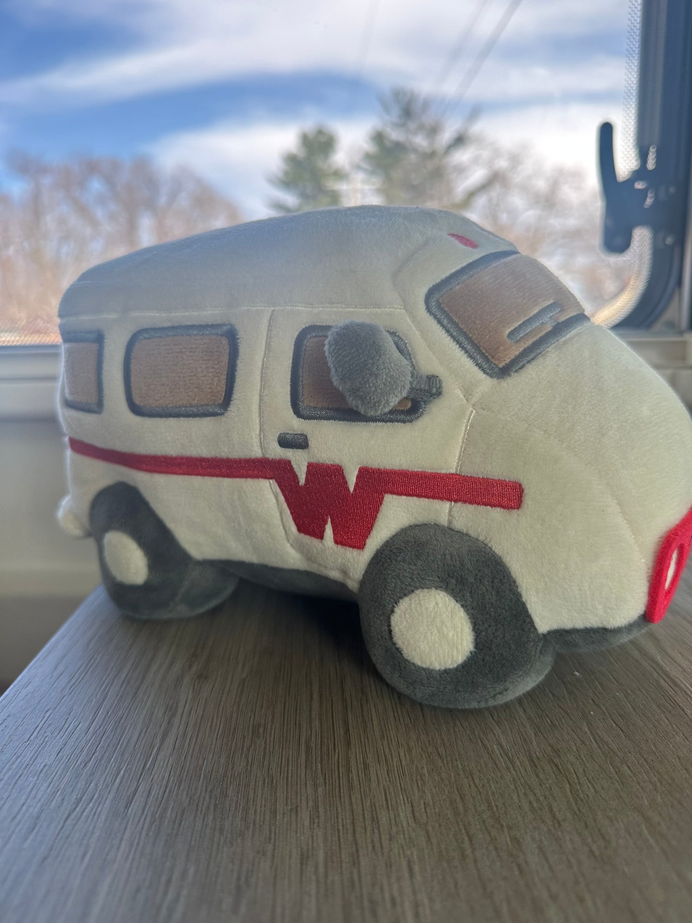 A cream plush RV toy with a red 'W' logo sits on a wood surface, against a blurred sky. Ready for your rv rental adventure!