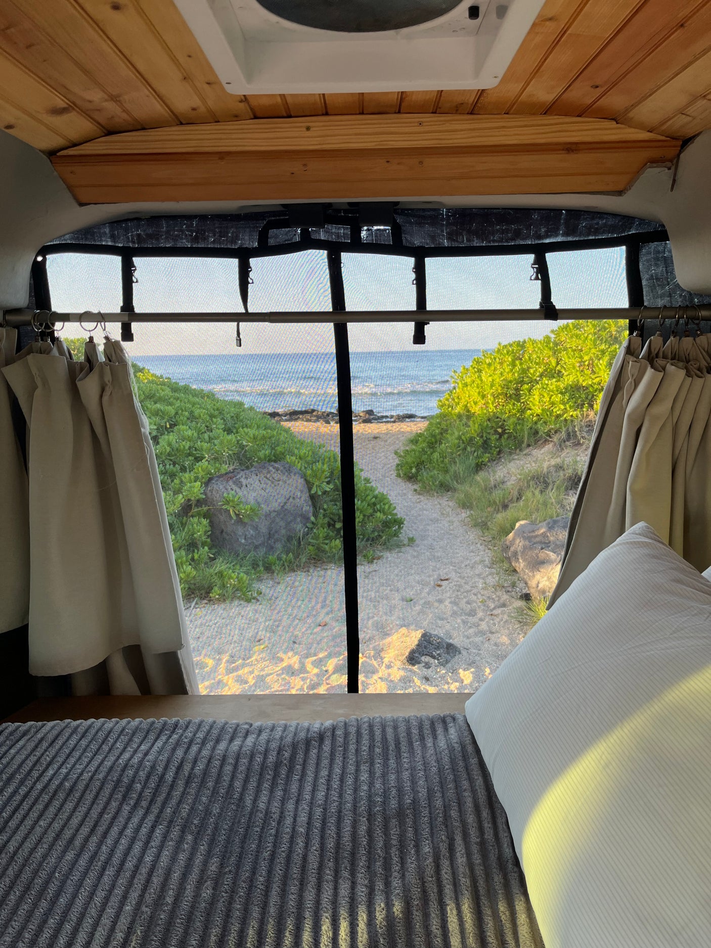 An RV rental's cozy interior, with a bed and curtains, overlooks a sandy beach path to the ocean. Perfect for scenic travel.