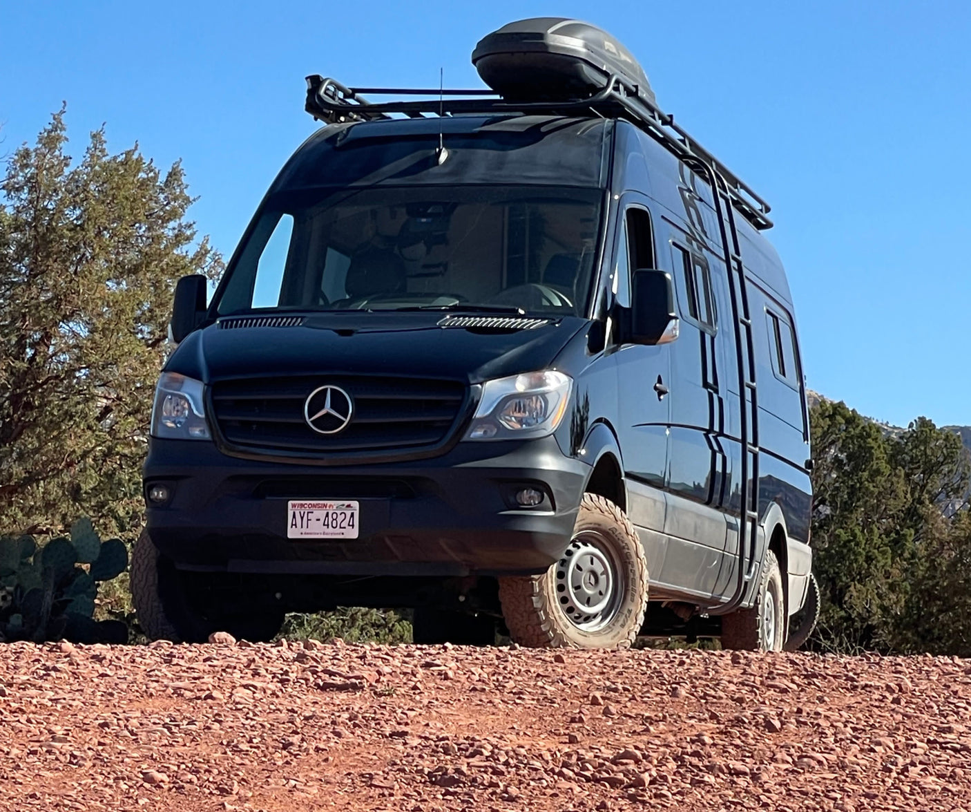 Black Mercedes Sprinter rv rental, equipped for adventure, parked on a reddish, rocky road under a clear sky.