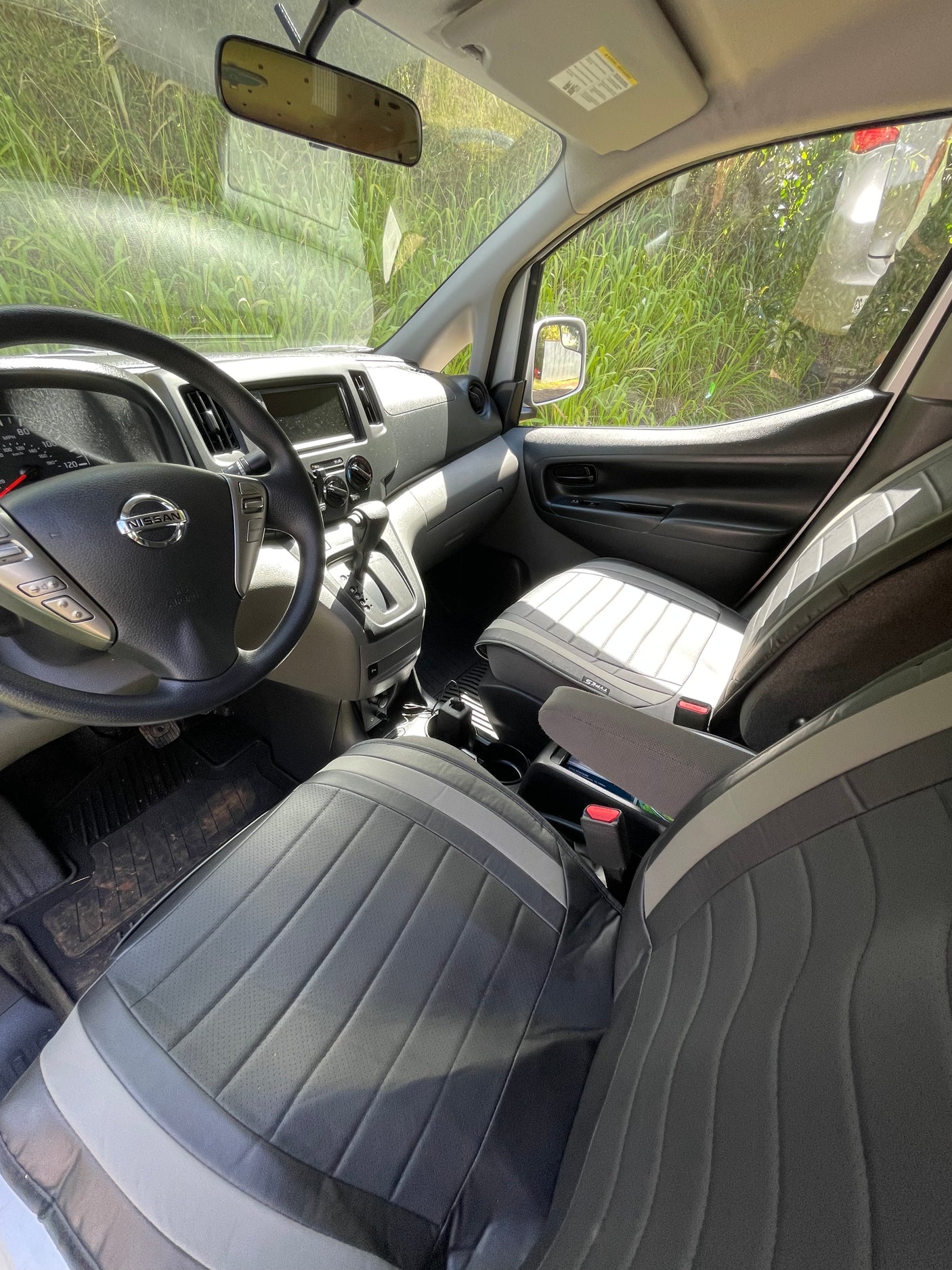 Interior of a Nissan van, perfect for your next RV rental adventure. Shows driver's cockpit, passenger seat, and green foliage outside.