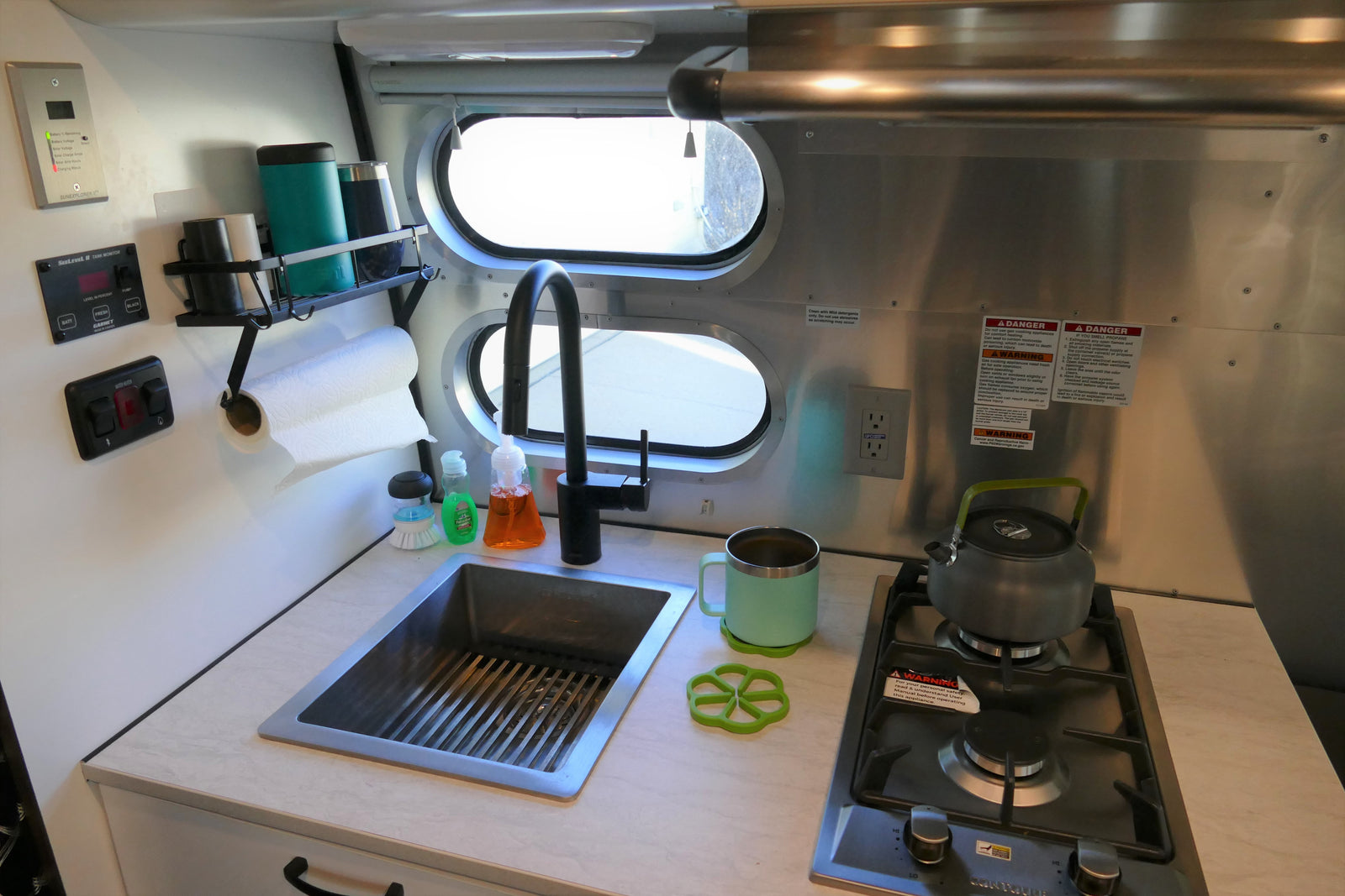 Compact kitchen in an RV rental, featuring a sink, two-burner stove, and monitoring displays for battery/water tanks.