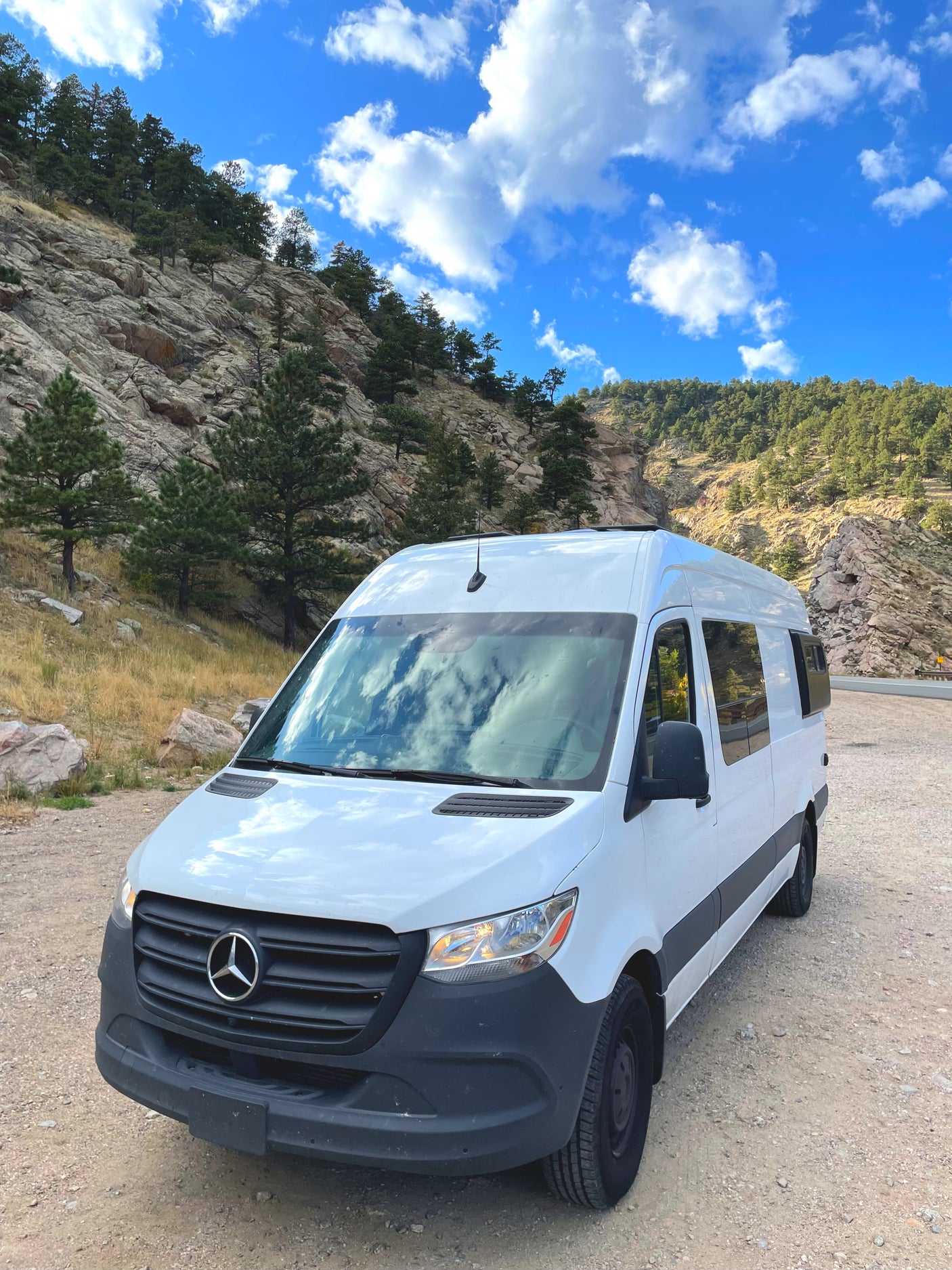 White RV rental van parked on a dirt road in scenic mountains under a bright blue sky with clouds.