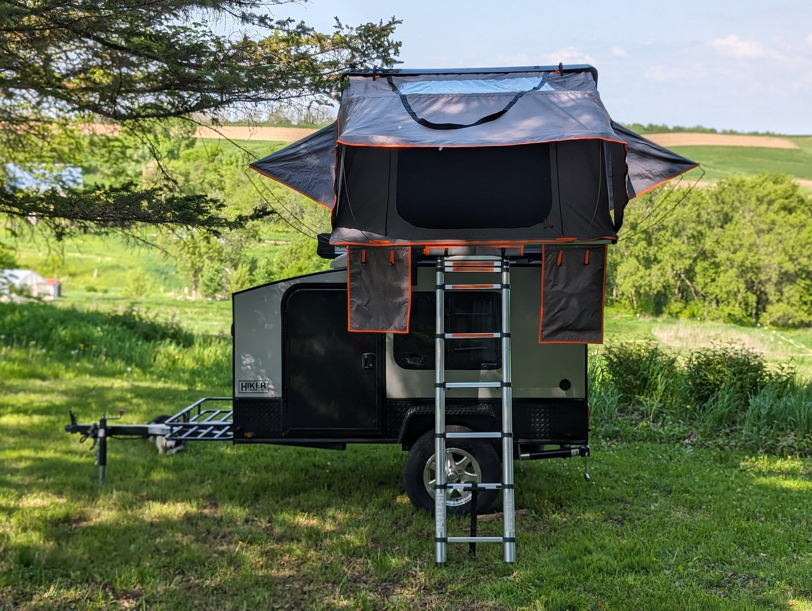 Rooftop tent camper trailer set up for a scenic outdoor adventure. Ideal for your next RV rental trip.|A white HIKER TRAILER RV rental emblem features a camper