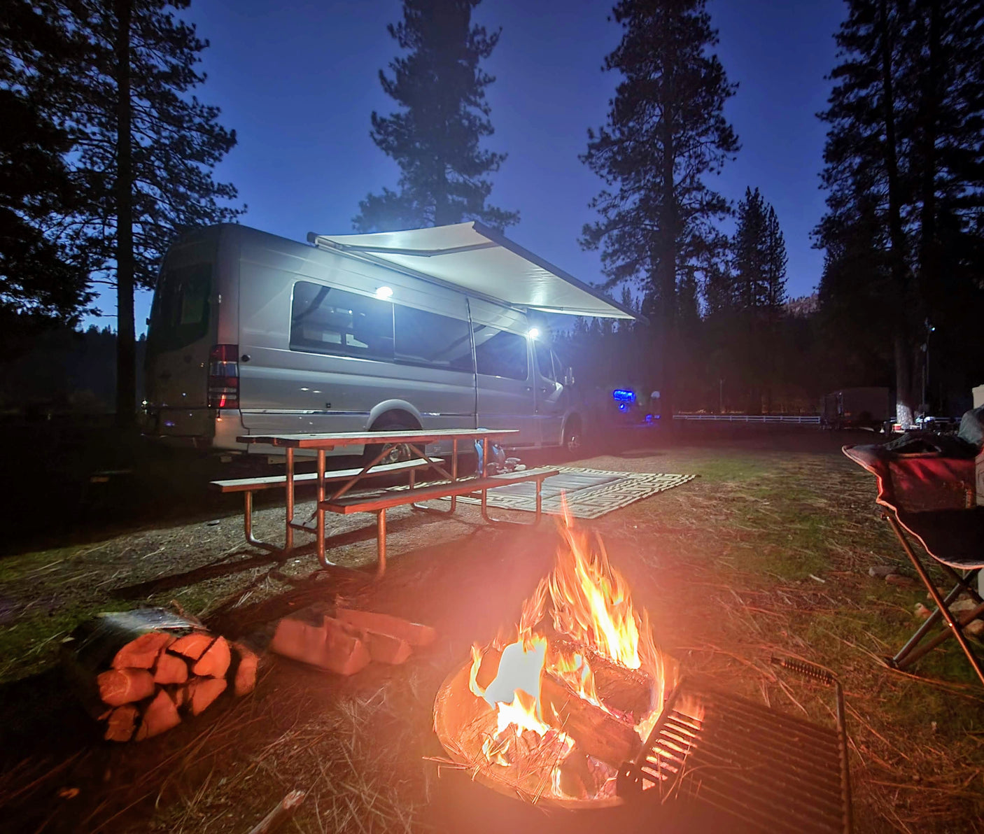 A cozy evening campsite featuring a white RV rental van with an extended awning, a crackling campfire, and a picnic table.