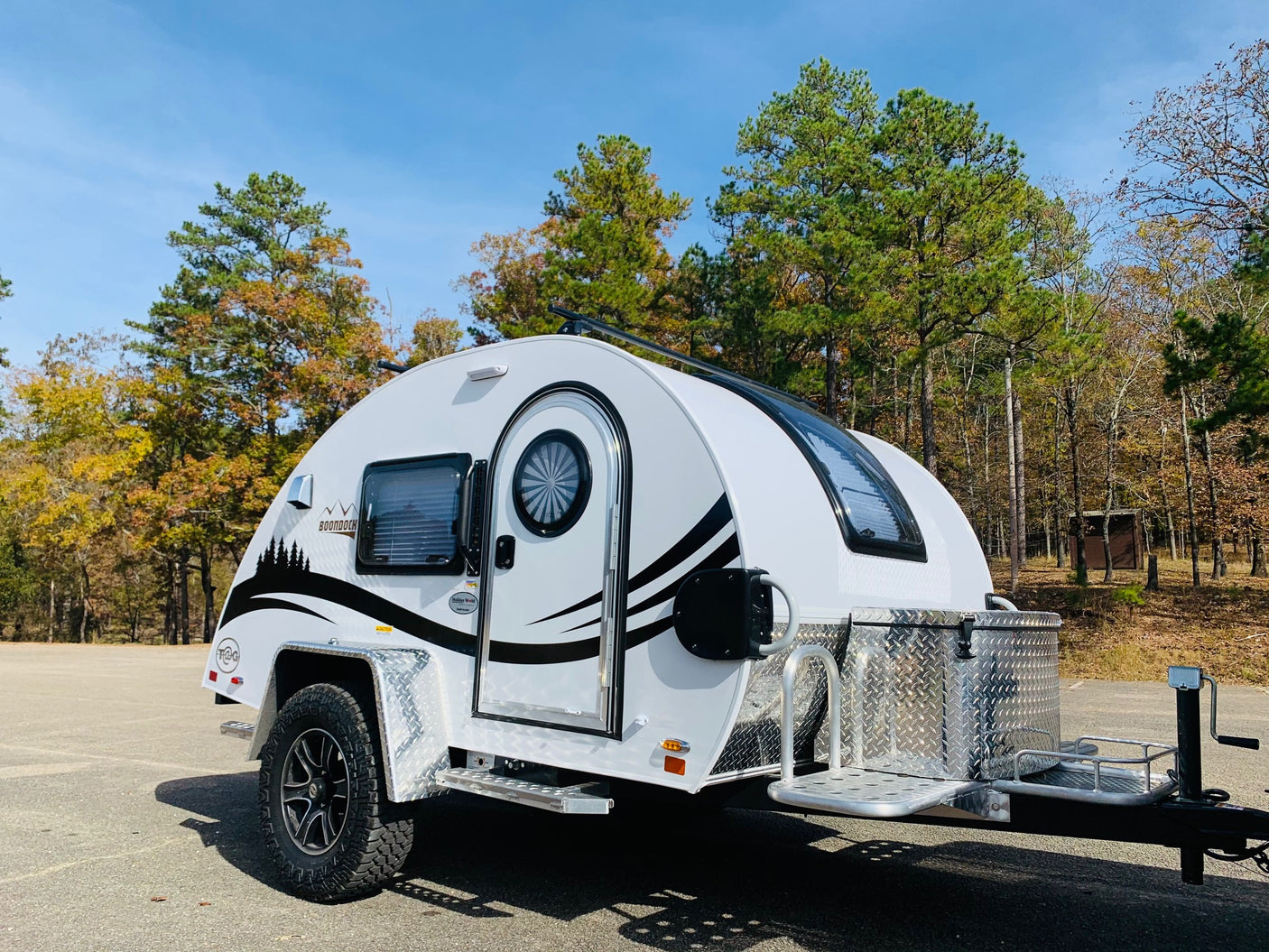 A white Boondock teardrop camper, an ideal RV rental, is parked on asphalt with colorful autumn trees.