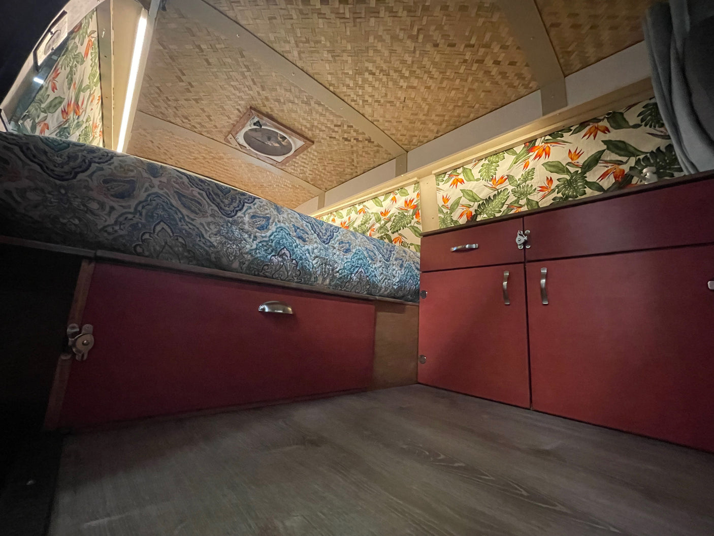 Cozy RV rental interior: blue patterned bed, red cabinets, tropical walls, woven ceiling, and wood floor.