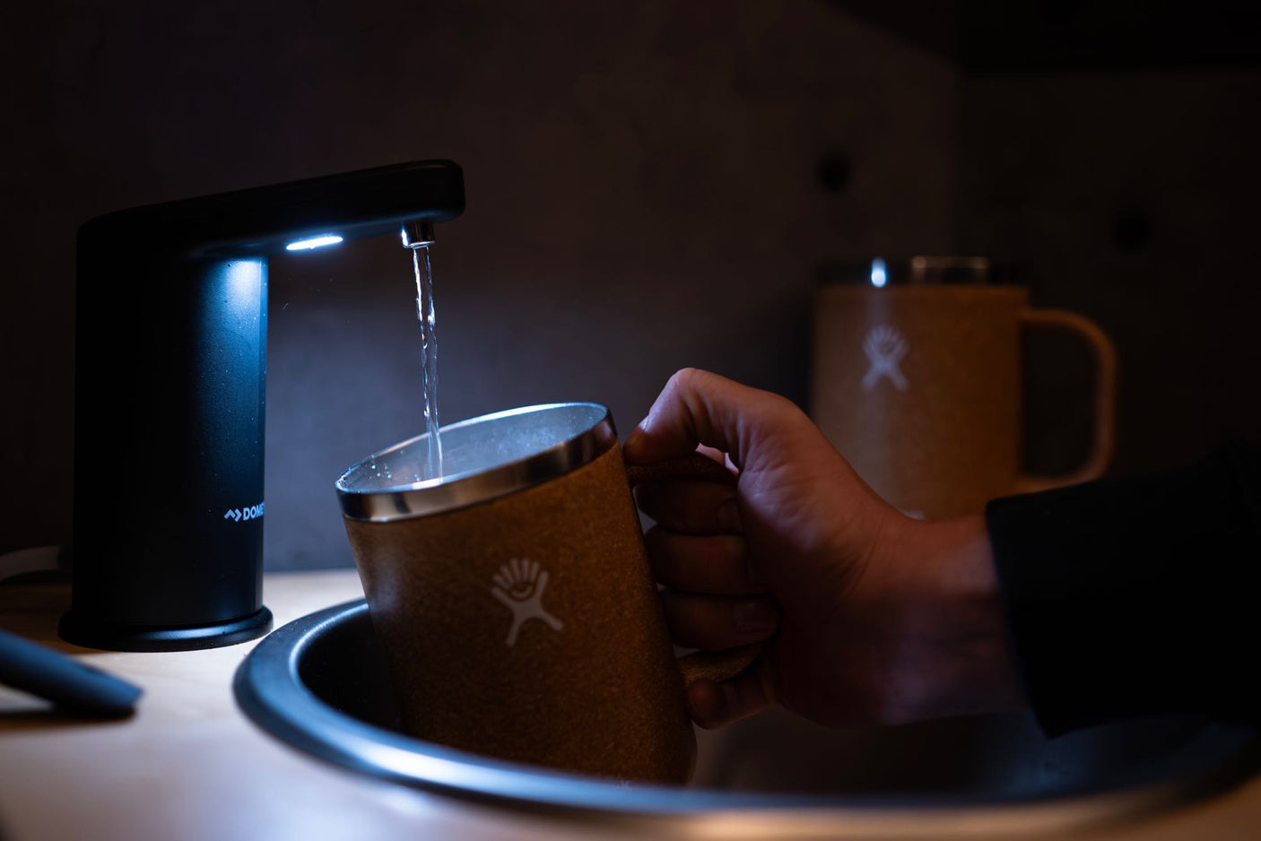 A hand fills a textured Hydro Flask mug from a Dometic water dispenser in an RV rental kitchen.