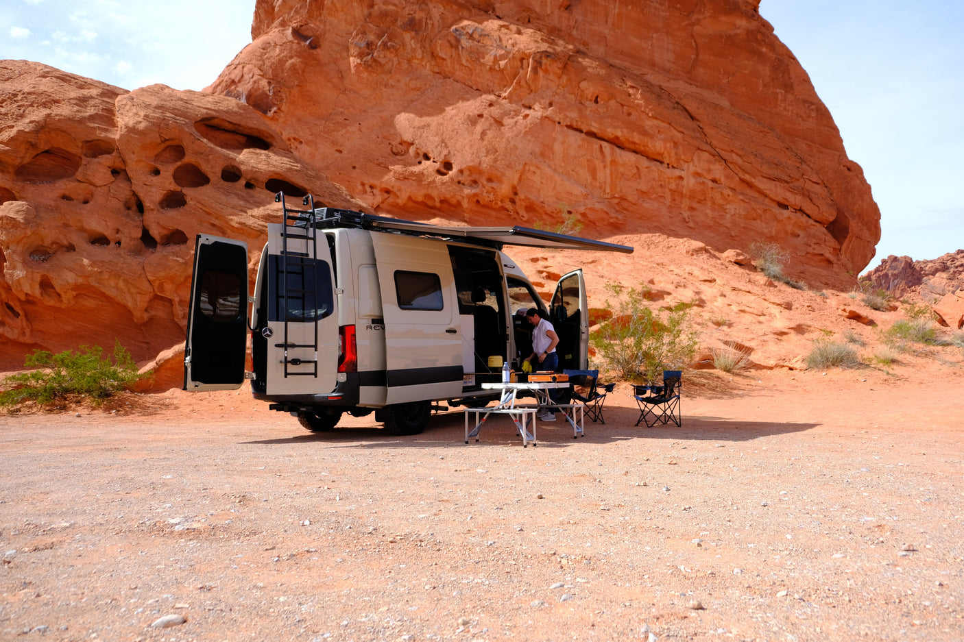 RV rental camper van set up for camping in a desert of red rock formations, with an awning, outdoor table, and chairs.