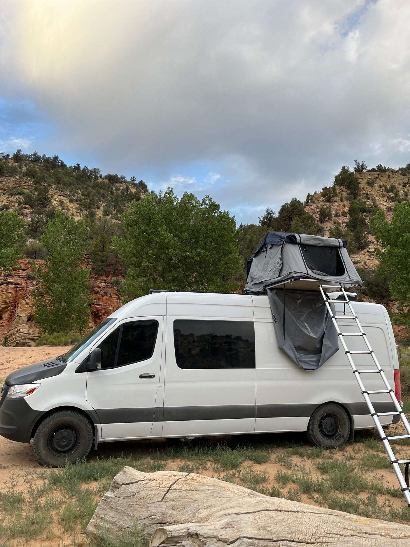 A white Sprinter RV rental with a rooftop tent and ladder deployed, parked for camping in a rocky, tree-lined landscape under cloudy skies.