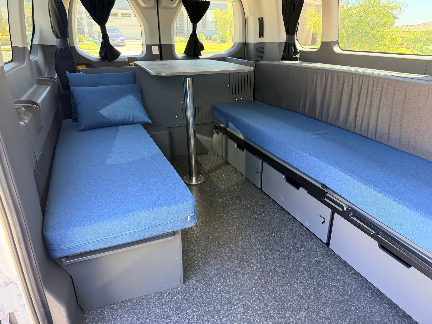 RV rental camper van interior: blue padded seating/beds, a central table, and under-bench storage drawers. Blackout curtains frame windows.