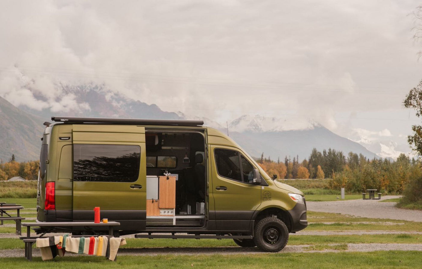 surrounded by vibrant fall foliage. Ideal for fall adventures.|Olive "Vanquest" camper van RV rental with extended awning on green grass. Scenic backdrop of trees and distant mountains.|Outdoor kitchen set up next to a bright green RV rental