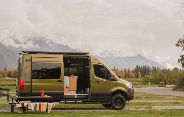 surrounded by vibrant fall foliage. Ideal for fall adventures.|Olive "Vanquest" camper van RV rental with extended awning on green grass. Scenic backdrop of trees and distant mountains.|Outdoor kitchen set up next to a bright green RV rental
