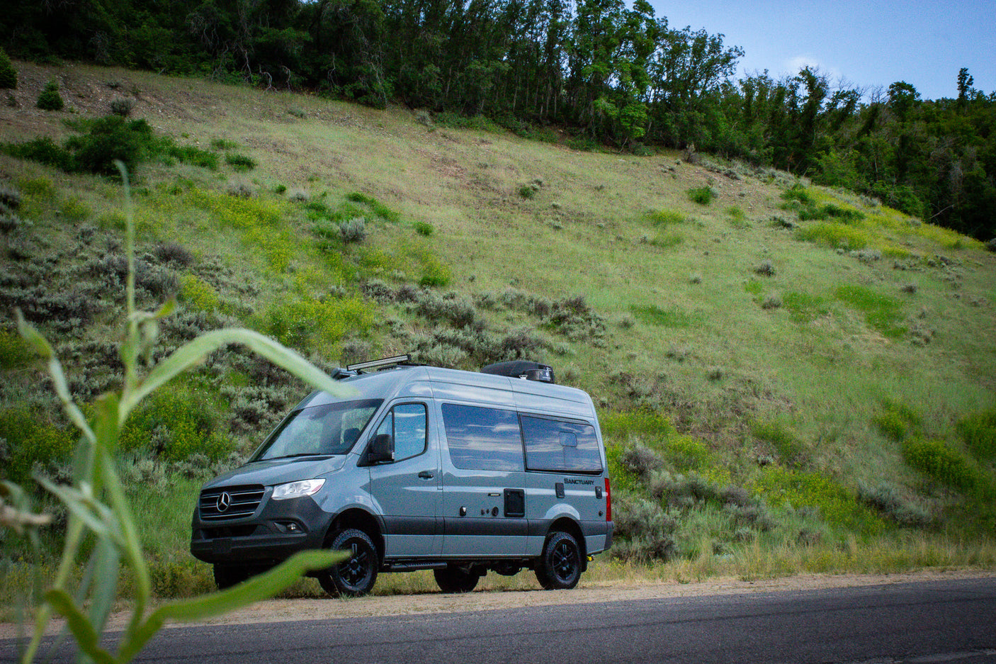 Grey Mercedes Sprinter RV rental parked on a roadside. Lush green hills & trees fill the background.