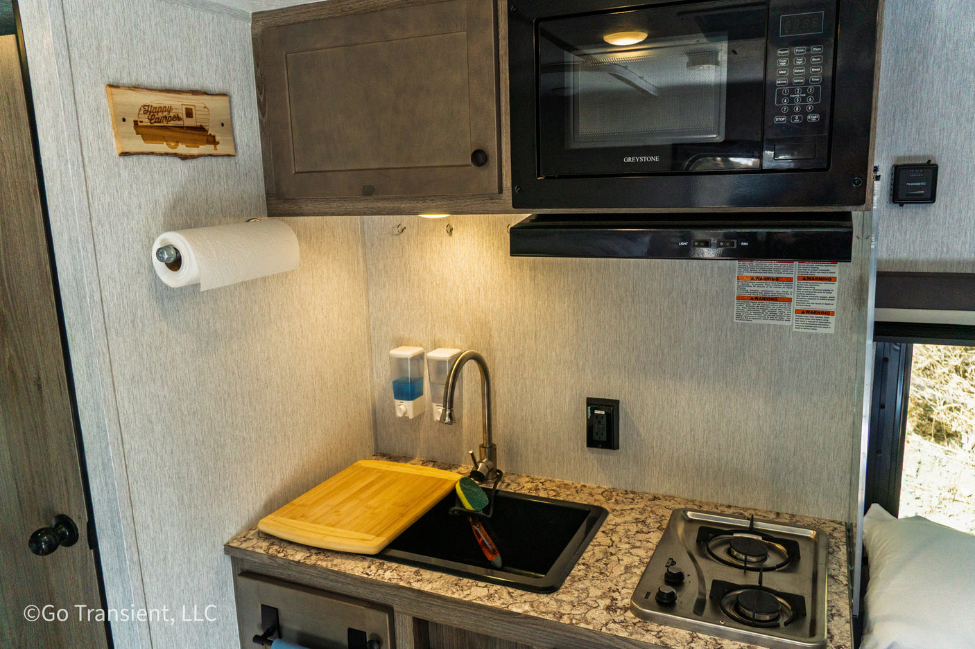 This compact RV rental kitchen features a microwave, stovetop, sink, paper towels, and a "Happy Camper" sign for your adventures.