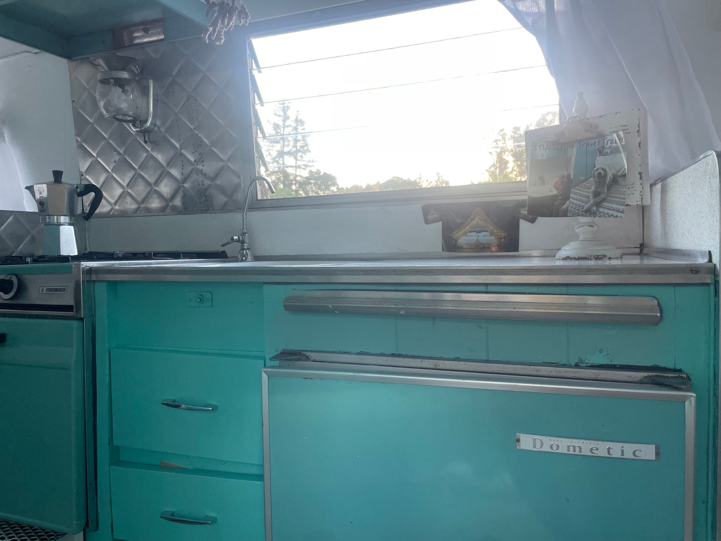 Vintage RV rental kitchen with teal cabinets, Magic Chef stove, Dometic fridge, and moka pot. Bright window view.