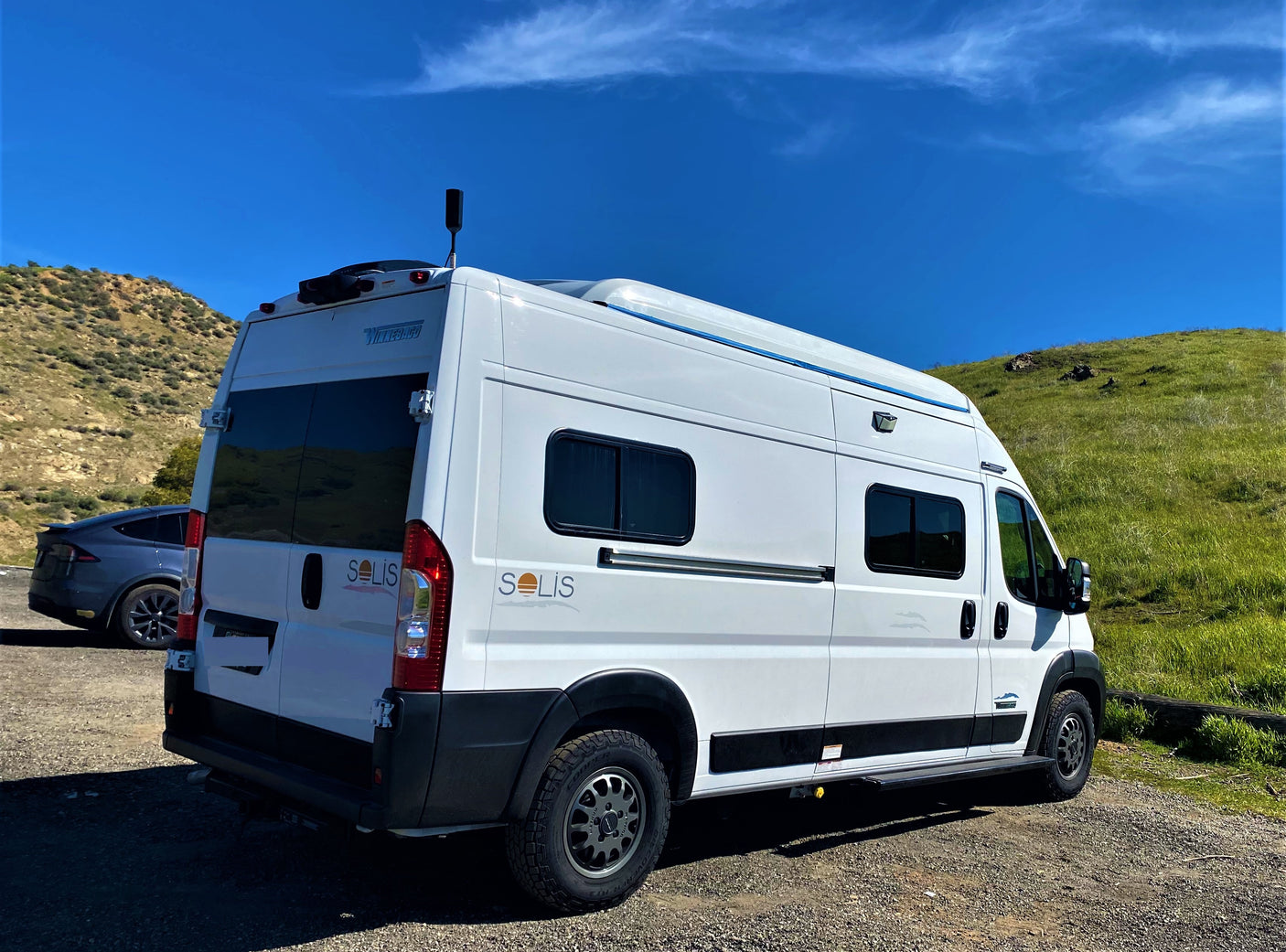 White Winnebago Solis camper van, great for an RV rental, parked on gravel by green hills under a bright blue sky. A Tesla is seen.