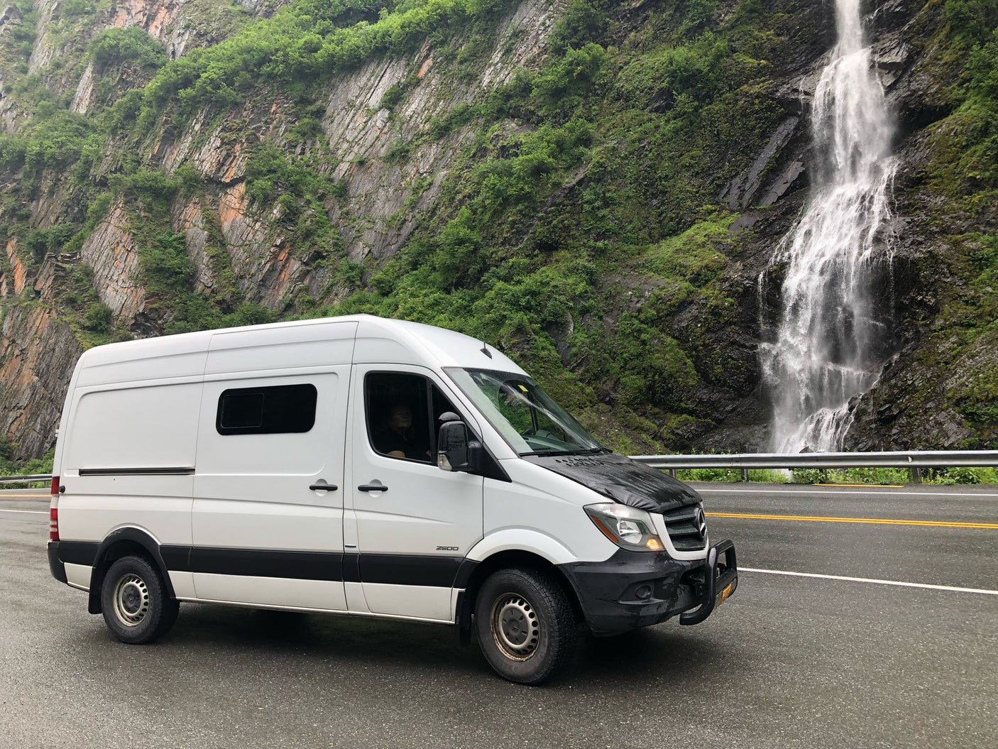 White Sprinter rv rental van beside a majestic waterfall and lush mountain on a rainy day.|An RV rental van parked beside a massive