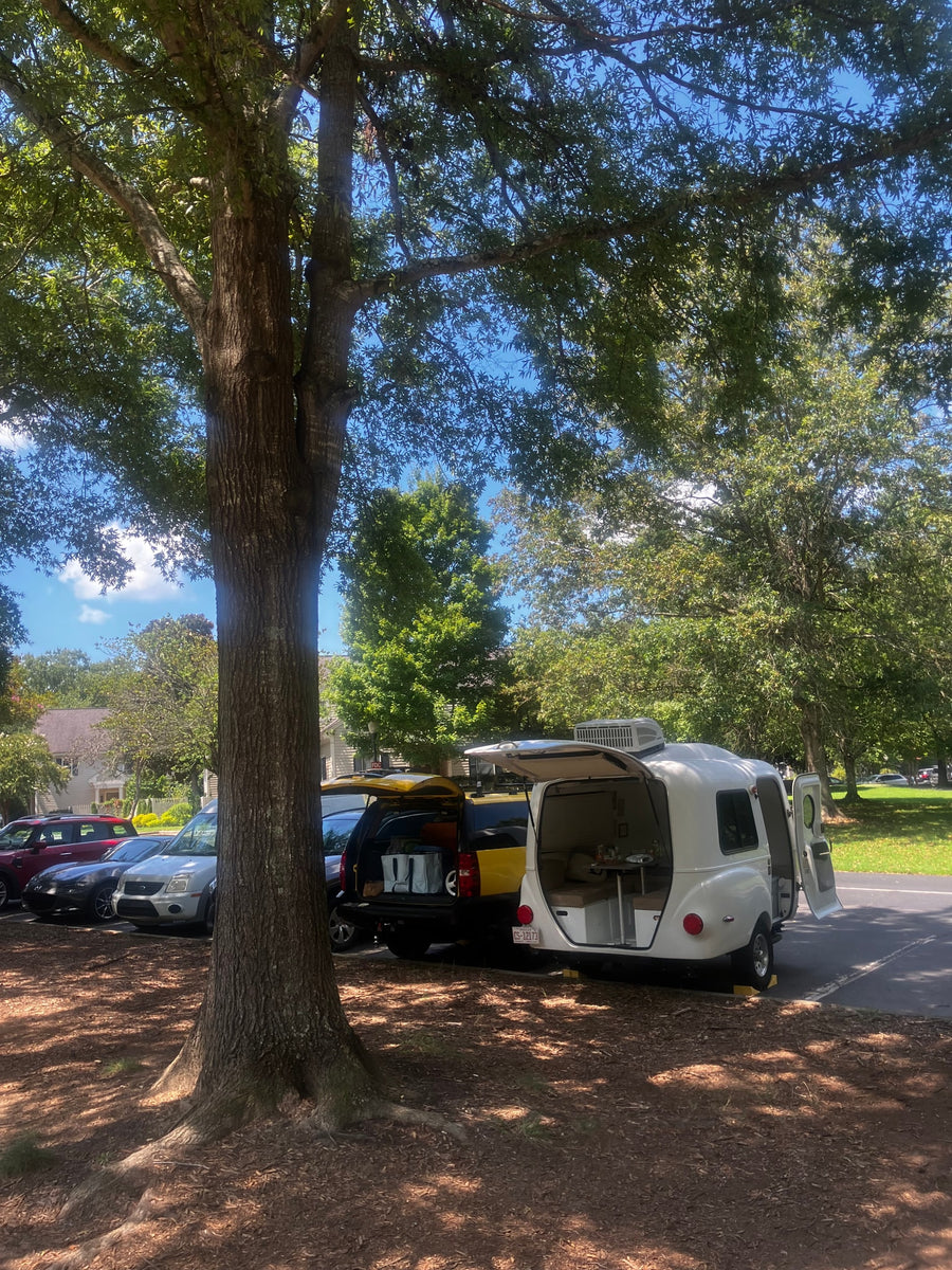 White teardrop RV rental parked under a large tree with its doors open, showing its interior, next to a yellow SUV.