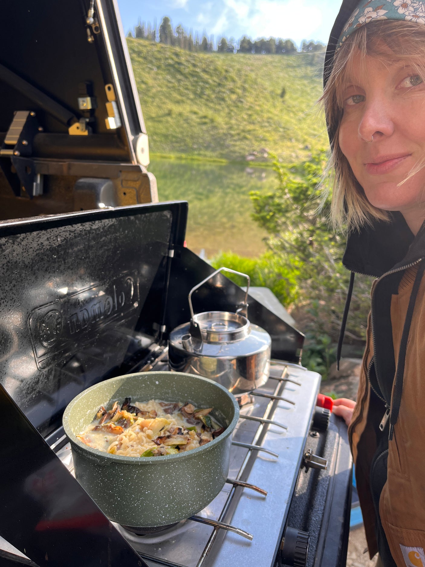 Woman cooking on a camping stove with a kettle by a lake, during an rv rental trip. Nature's beauty enhances the outdoor meal.