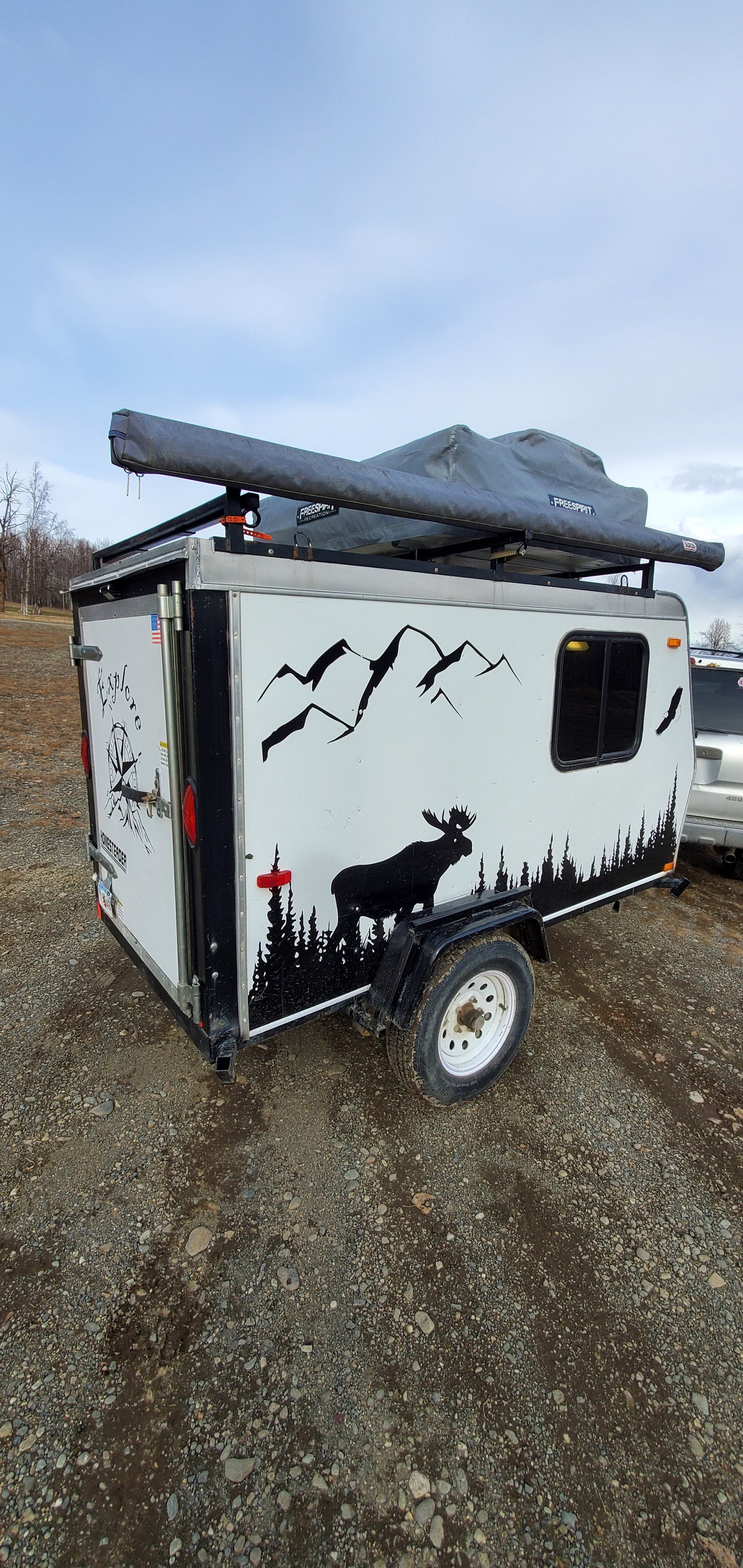 Compact adventure RV rental: A white camping trailer with nature decals and rooftop tent, parked on gravel, perfect for outdoor exploration.