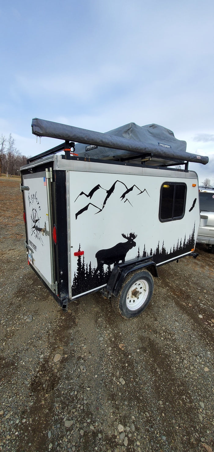 Compact adventure RV rental: A white camping trailer with nature decals and rooftop tent, parked on gravel, perfect for outdoor exploration.