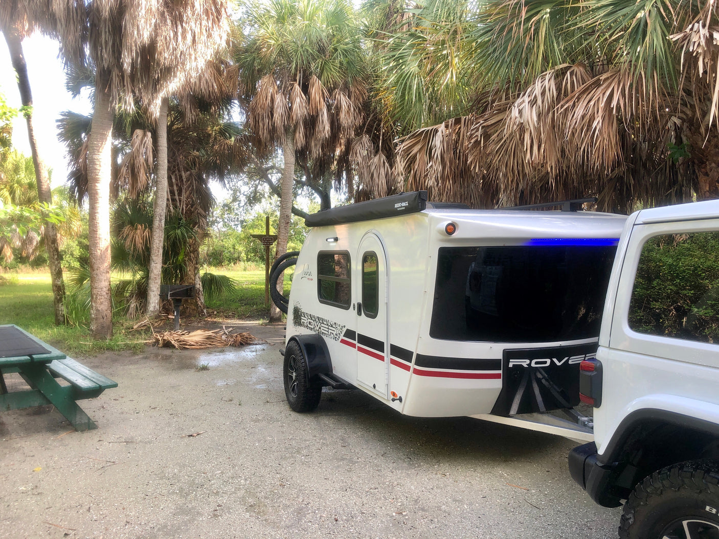 Your rv rental adventure awaits! A white travel trailer hitched to a Jeep at a palm tree campsite with a picnic table.