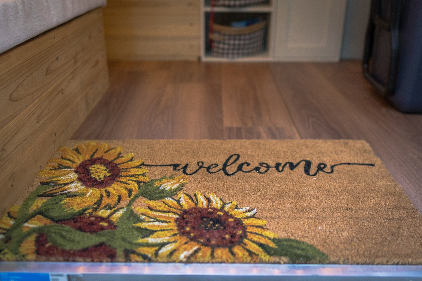A 'welcome' mat with sunflowers adds a cozy touch to an RV rental interior, featuring wood floors and built-in storage.
