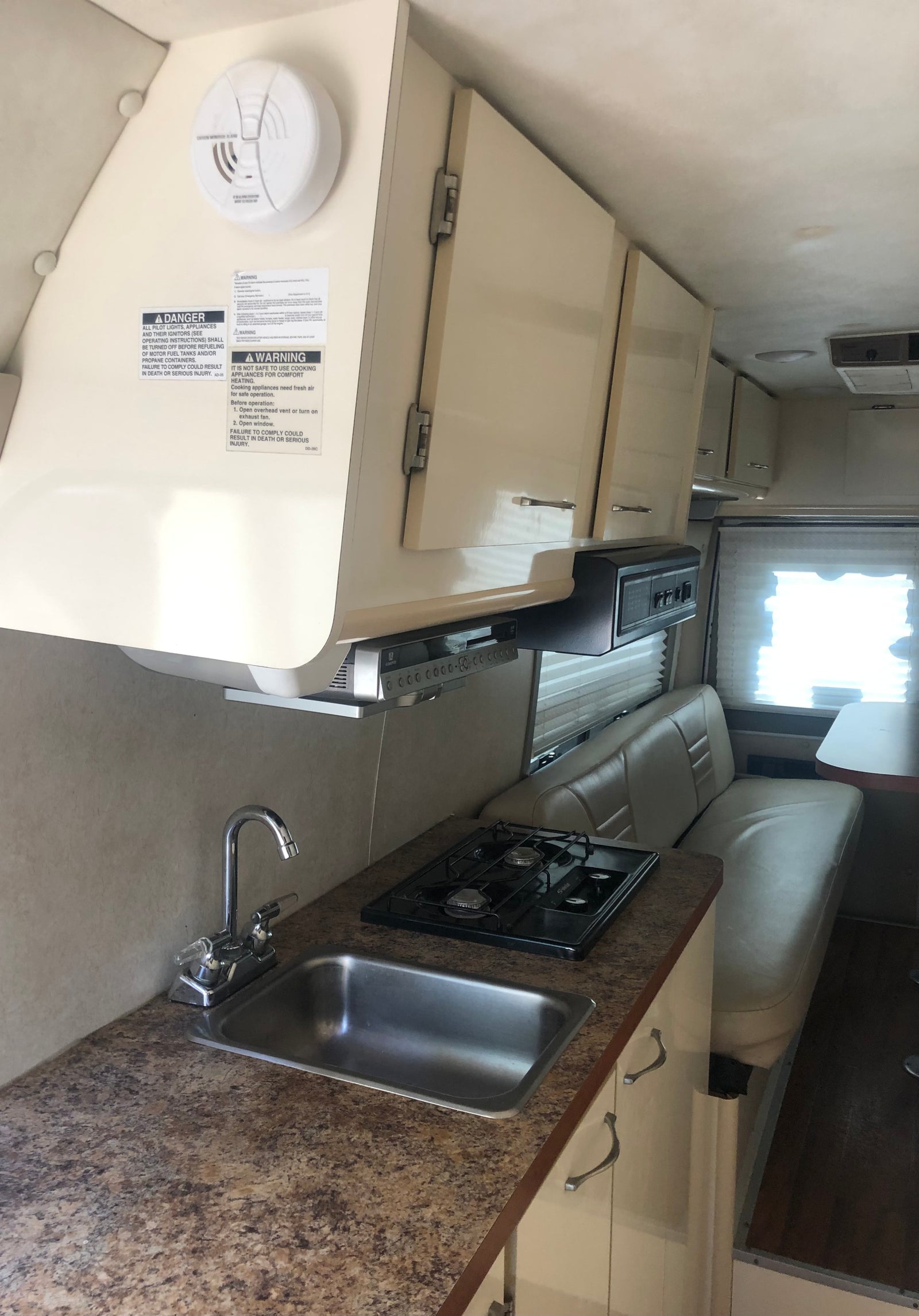 Interior view of an RV rental showing a small kitchen area with sink, 2-burner stove, overhead cabinets, and dinette.