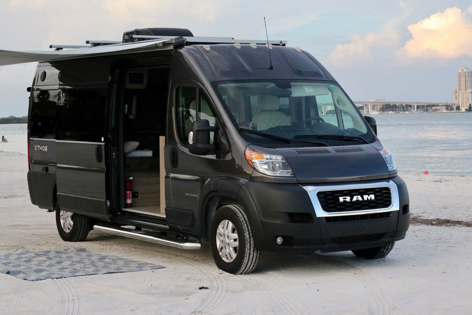 Dark gray Ram camper van, ideal for rv rental, parked on a beach by the water. Awning deployed, side door open.