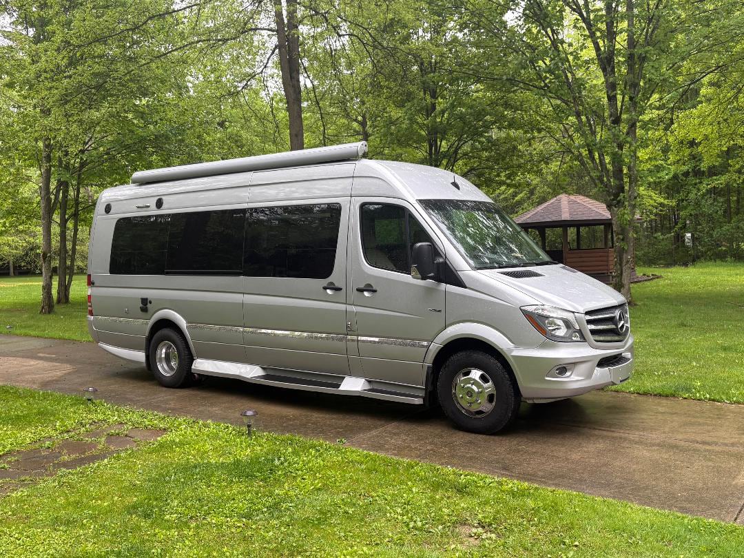is parked on a wet paved path surrounded by lush trees.|Silver Mercedes Sprinter RV rental