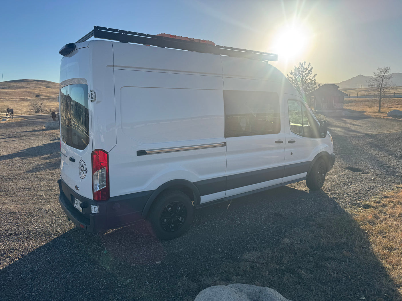 White RV rental camper van parked on gravel, strong sunflare, roof rack, hills, and building in background at sunset.