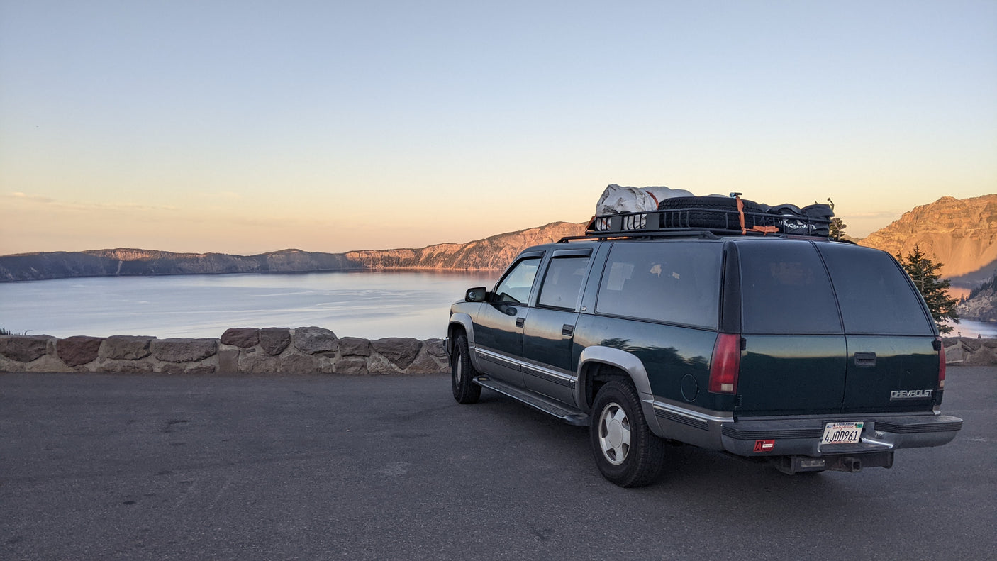 This dark green Chevrolet Suburban, outfitted like an RV rental, is parked overlooking a vast lake and mountains at sunrise.