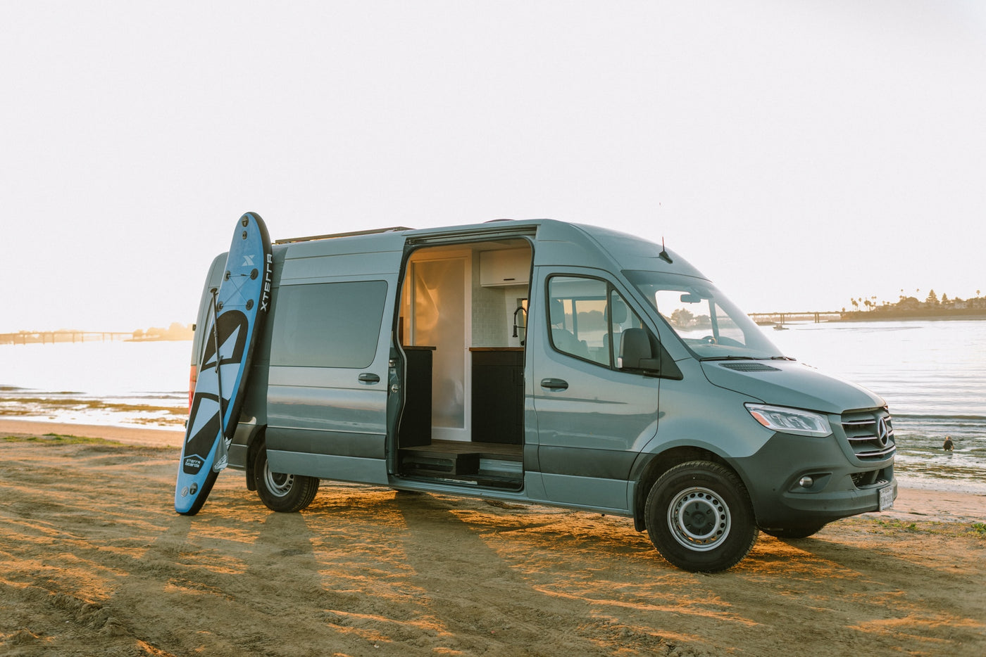This custom Mercedes Sprinter RV rental parked by the bay with an open side door reveals a full camper interior and paddleboard. — photo 38