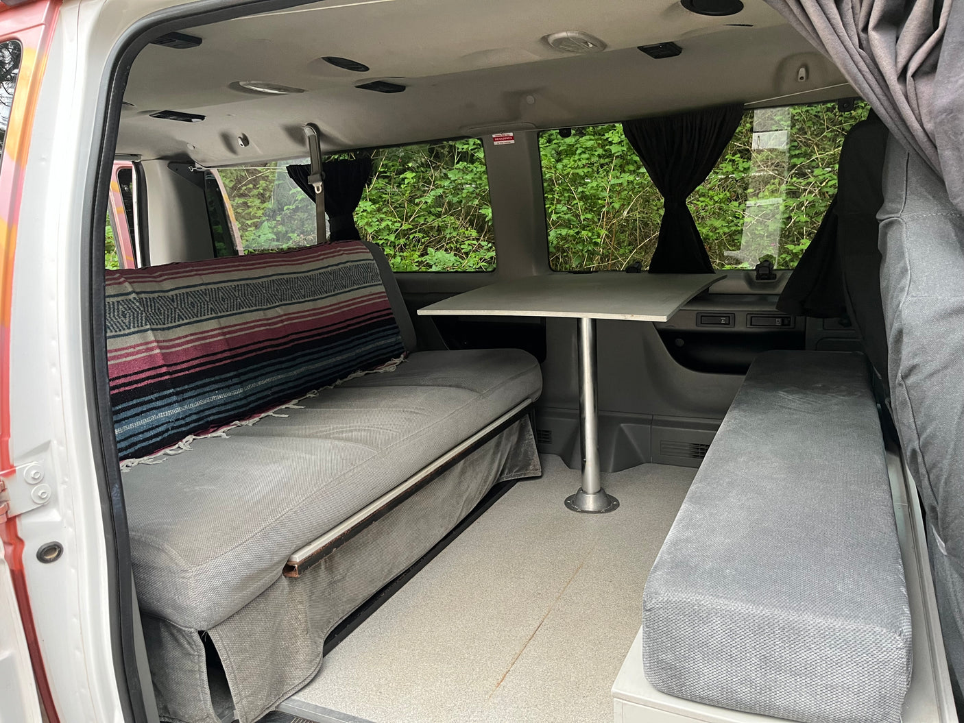 Cozy RV rental interior with bench seating and a dining table, perfect for travel adventures. Windows show lush greenery.