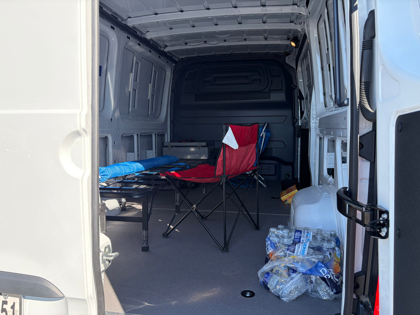 RV rental cargo van interior: cot, red chair, water bottles, and storage box on a carpeted floor.