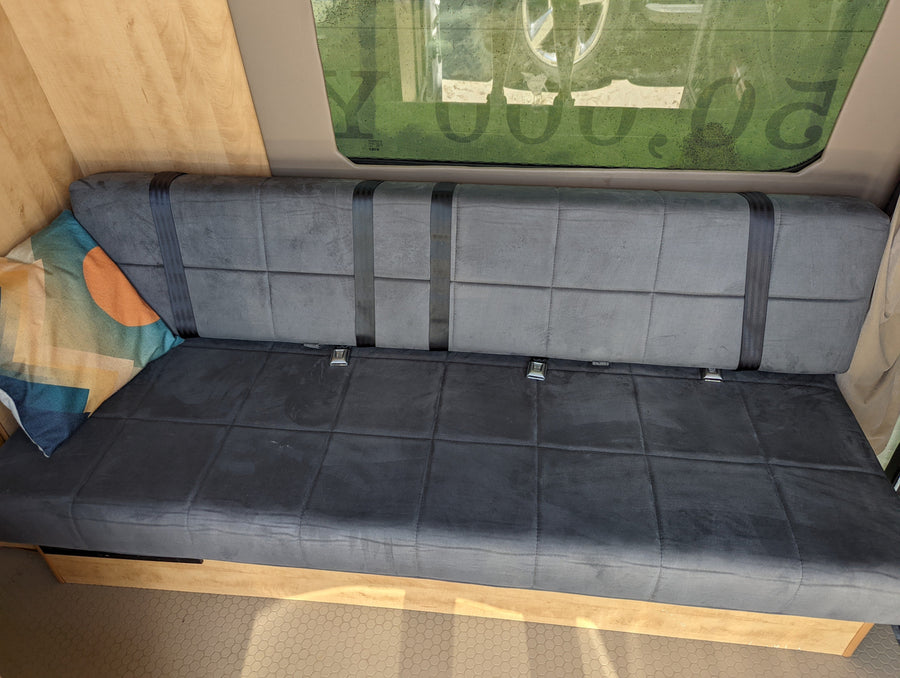 RV rental interior shows a gray bench seat with black seatbelts, a patterned pillow, and wooden walls next to a window.