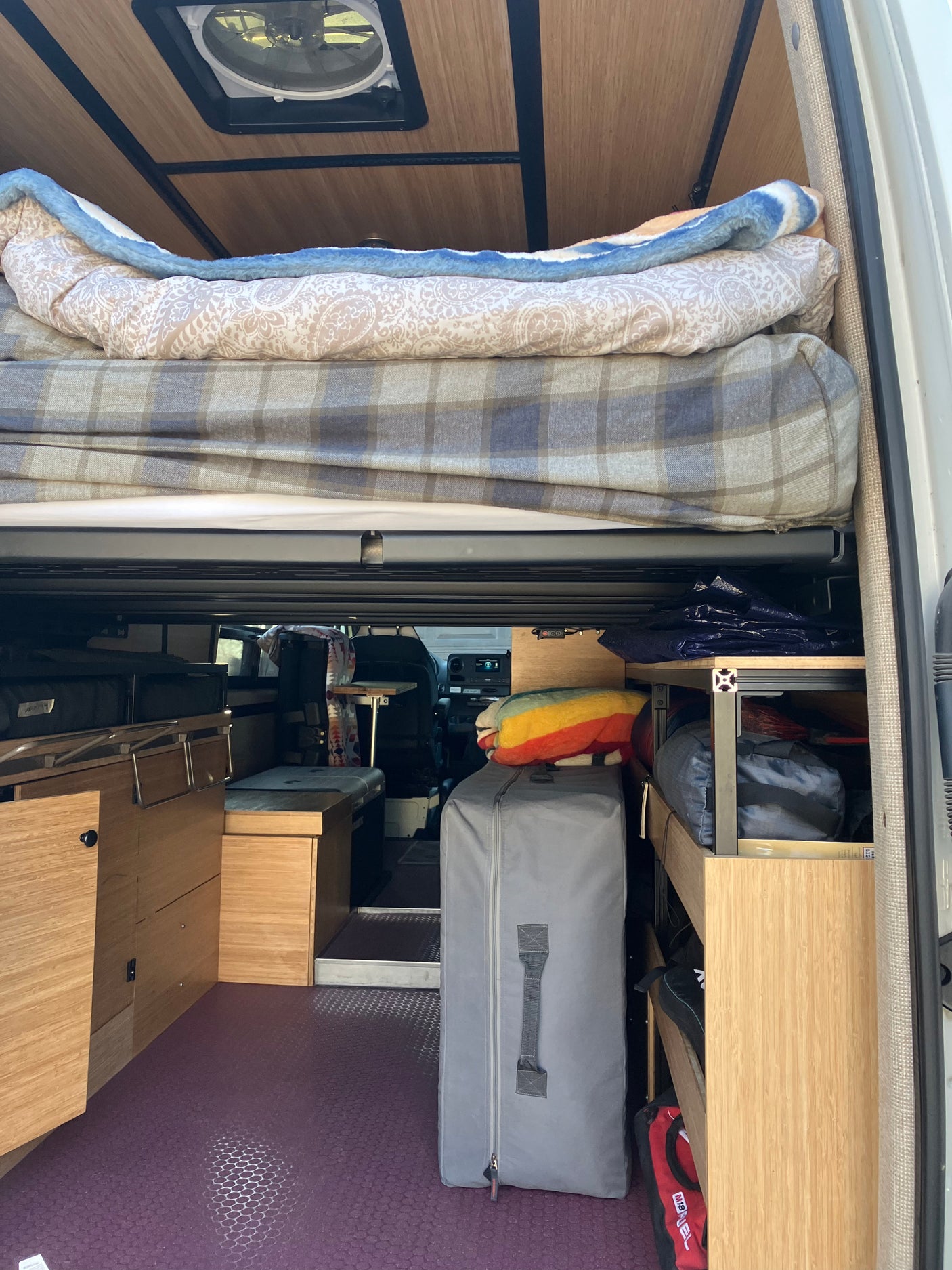 An rv rental camper van's interior, showing a raised bed, wooden cabinets, organized storage, and a roof vent.