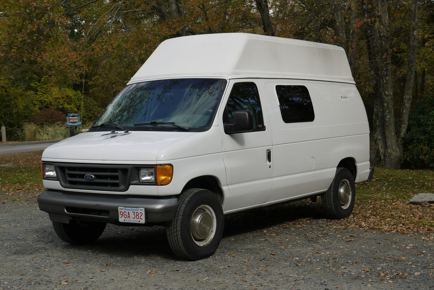 parked on a gravel lot amidst autumn trees.|White high-top Ford RV rental van parked on gravel in autumn