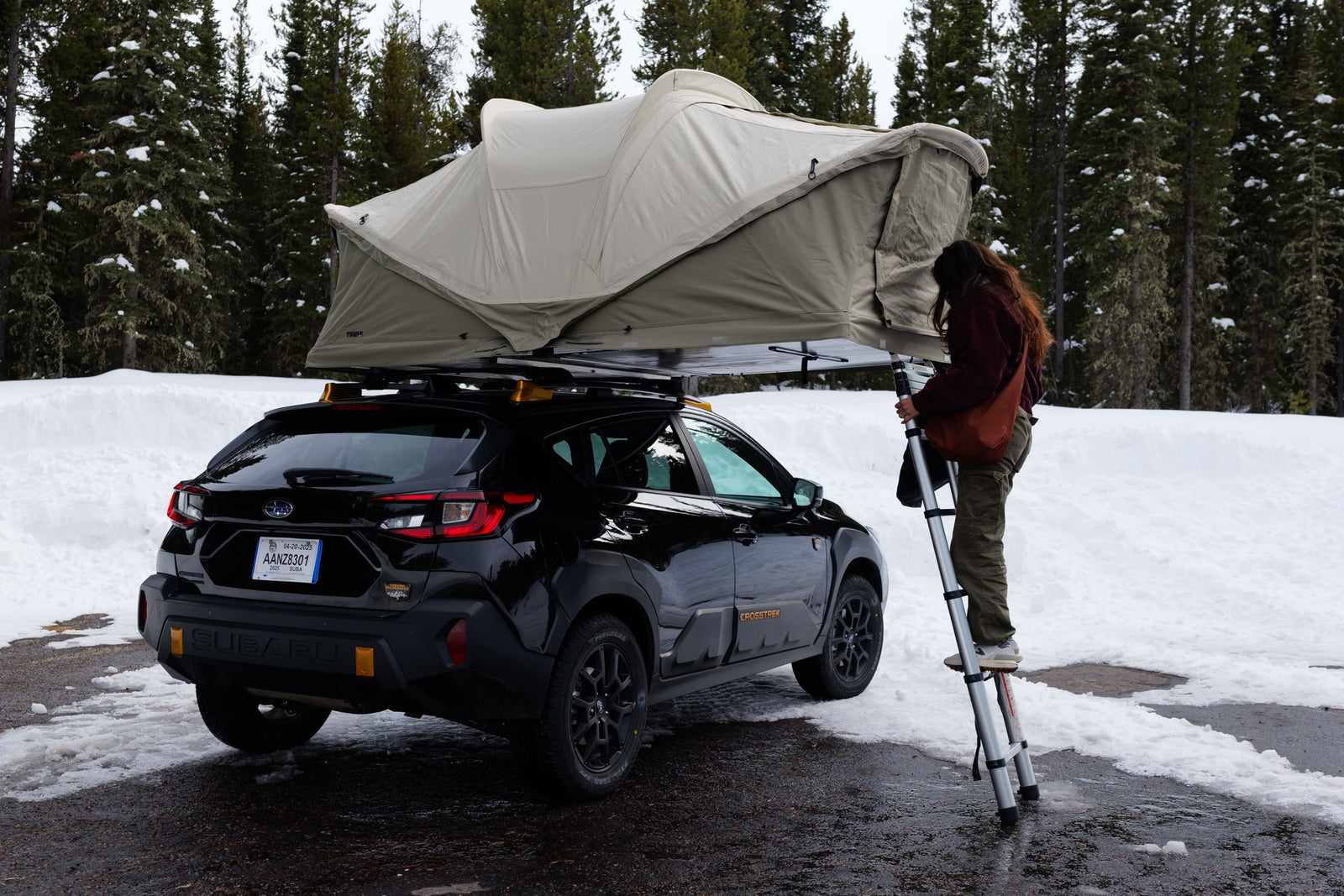 Person setting up Thule roof-top tent on black Subaru Crosstrek amidst snowy trees, ideal for an RV rental alternative.