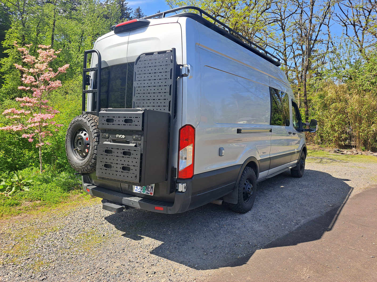 Silver camper van, equipped for adventure, seen from rear-right. Ideal for RV rental. Features roof rack, ladder, and rear storage.