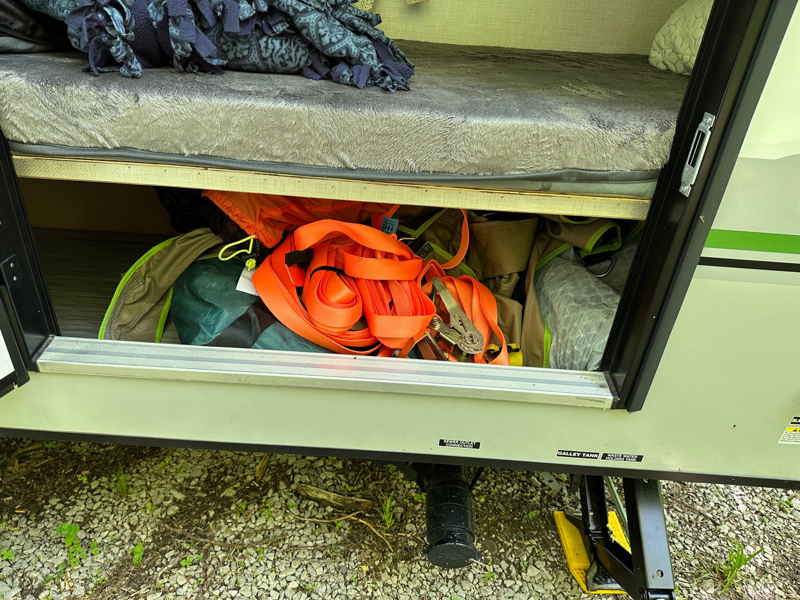 RV rental under-bed storage filled with camping gear. Exterior reveals wastewater tanks and a stabilizer jack.