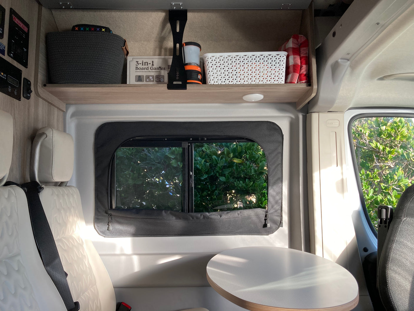 Inside an RV rental: compact living area with comfy seats, a table, shaded window, and overhead storage for games & essentials.