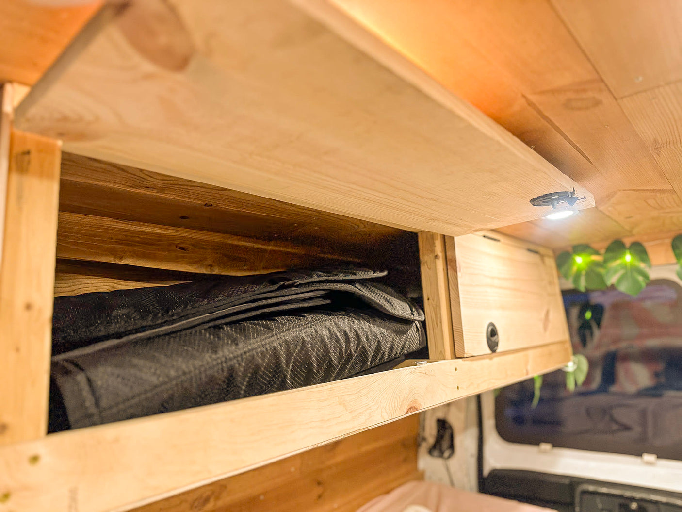 Overhead wood storage in an RV rental, open with folded black padded covers inside. An LED light shines above.