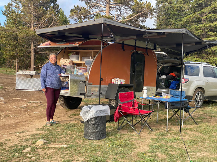 Woman smiles next to her orange tear-drop camper with an awning setup, perfect for an RV rental getaway in nature.
