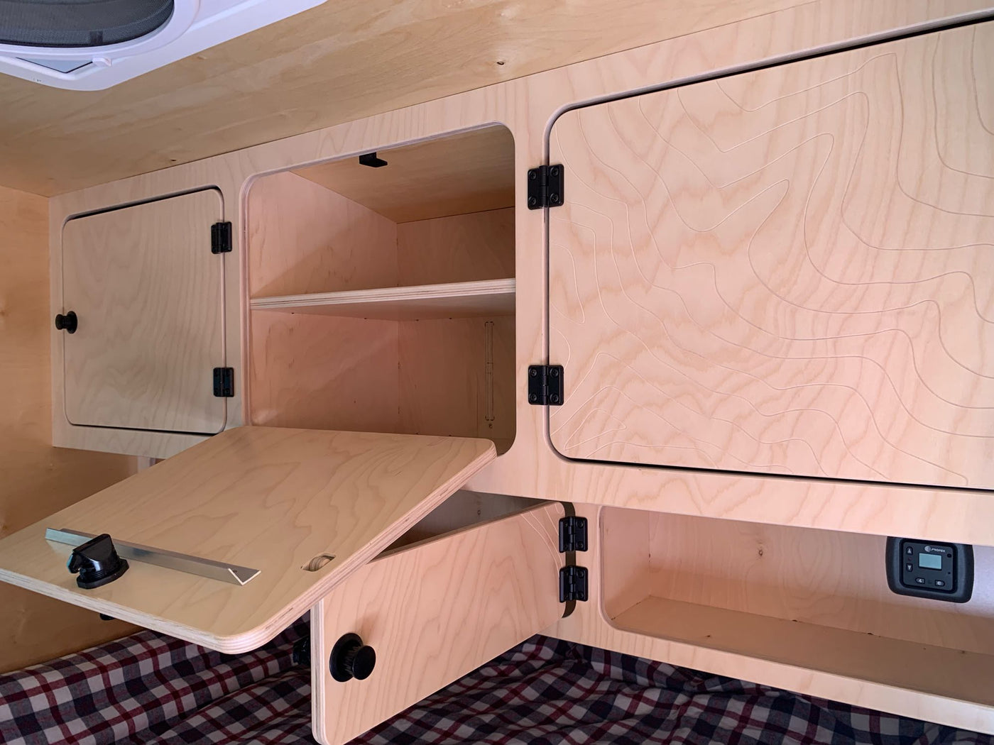 and a climate control unit over plaid bedding.|Custom birch plywood interior of an RV rental: built-in cabinets with topo map designs — photo 5