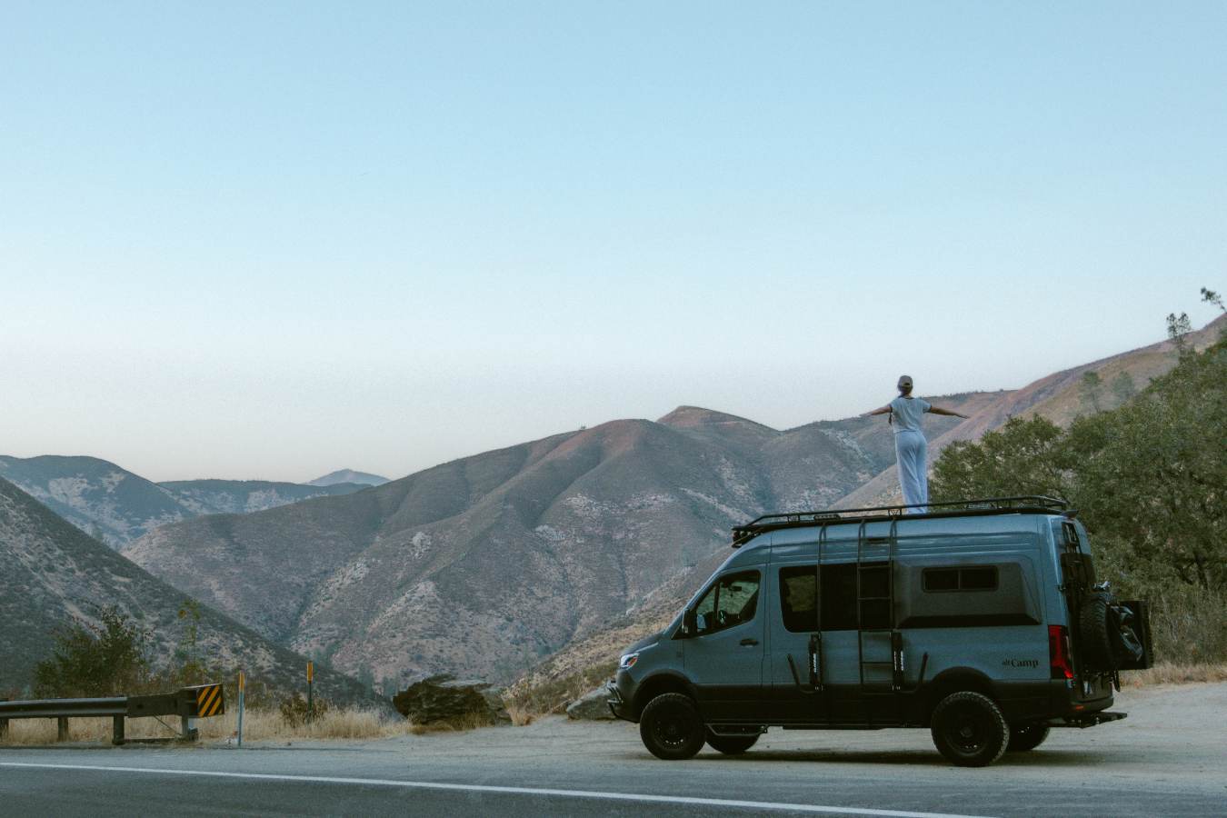 Embrace adventure! This RV rental offers stunning mountain views. A person enjoys van life freedom, standing on the van's roof.