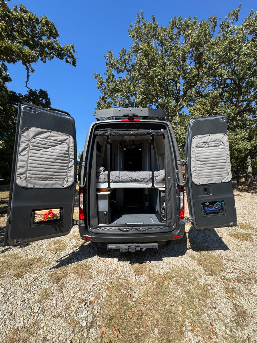 Spacious rv rental van ready for adventure. Open doors reveal bed & gear, parked on gravel under sunny skies.