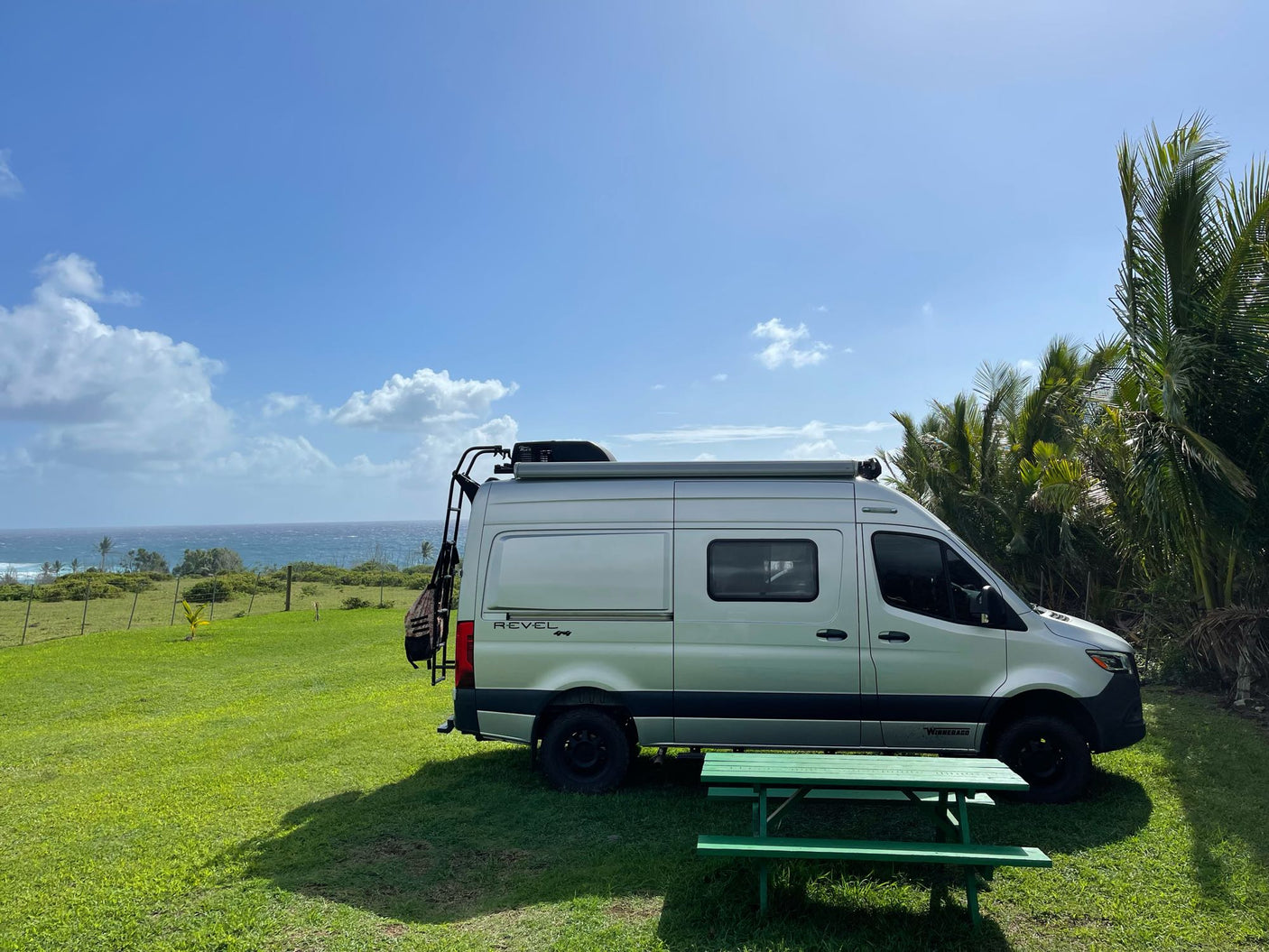 Silver Winnebago Revel 4x4 rv rental on green lawn, overlooking ocean, with palm trees & picnic table.