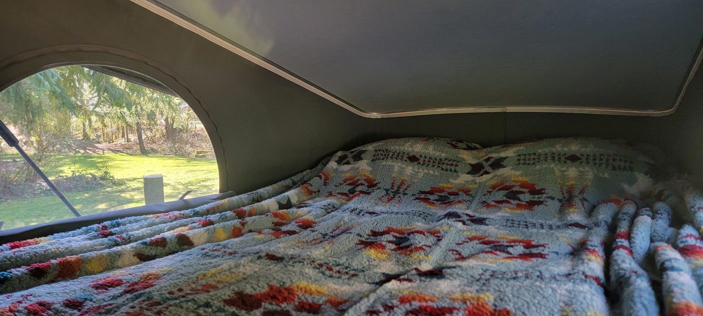 Cozy RV rental bed with a colorful, patterned blanket, featuring a scenic nature view through the pop-top window.